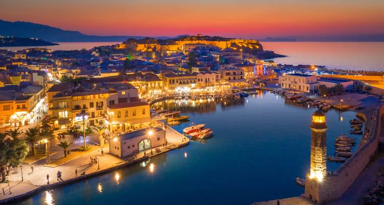Lebendiger Sonnenuntergang über einer erleuchteten Hafenstadt mit Spiegelungen auf dem Wasser.