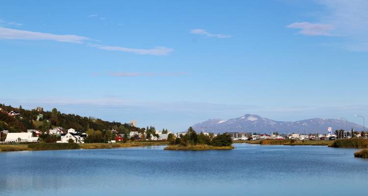 Quiet lake with distant town and mountains under a clear blue Icelandic sky.