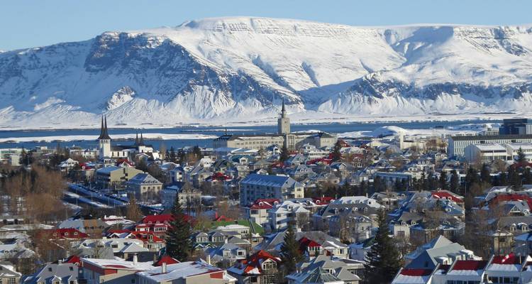 Panoramablick auf Reykjaviks farbenfrohe Dächer vor dem Hintergrund schneebedeckter Berge mit dem hohen Turm der Hallgrimskirkja-Kirche.