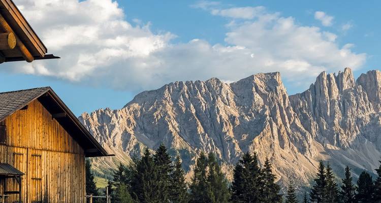 Un granero rústico de madera se asienta bajo imponentes picos escarpados de las Dolomitas y bosque verde.