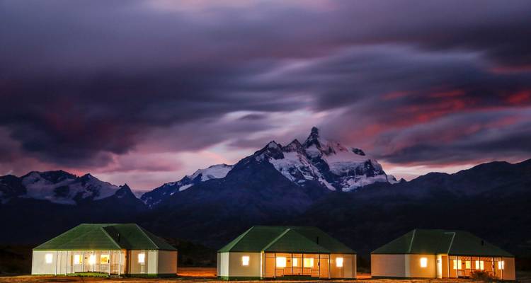 Drie moderne lodge-gebouwen liggen voor met sneeuw bedekte bergen onder een levendige zonsonderganghemel