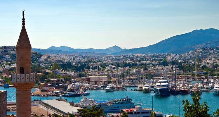Harbour of Bodrum with yachts, whitewashed town and surrounding hills seen past a mosque minaret.