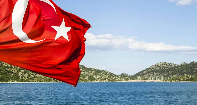 Turkish flag waving over a tranquil blue bay with green hills beyond.