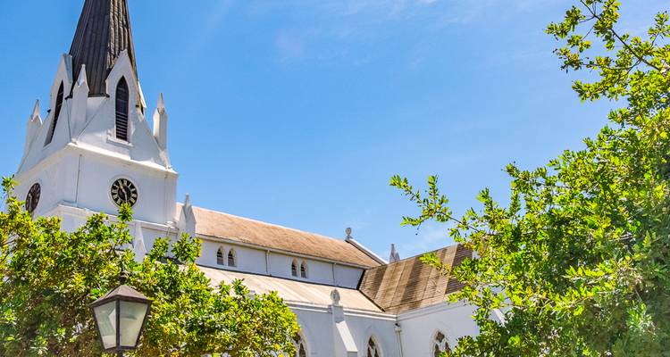 Weiß getünchte Kirche mit spitzem Kirchturm ragt über belaubte Bäume unter einem strahlend blauen Himmel.