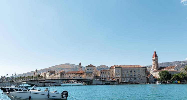 La histórica orilla de Trogir y su puente de arco de piedra visto a través del agua azul y tranquila con una pequeña lancha motora en primer plano.