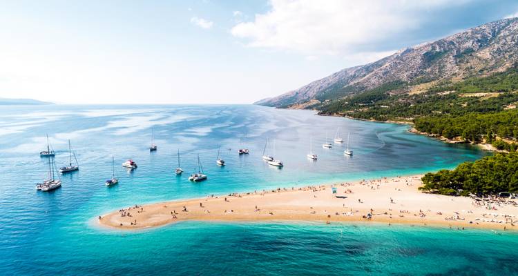 Vista aérea de botes anclados frente a la lengua de arena de Zlatni Rat donde las aguas turquesas poco profundas se encuentran con el canal azul más profundo, enmarcado por montañas y bosque de pinos.