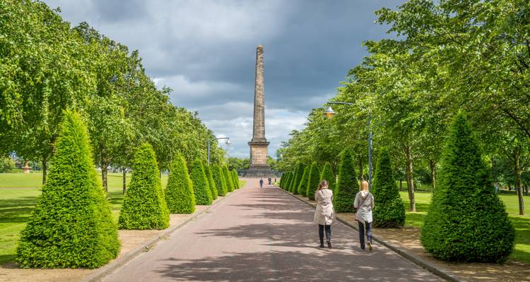 Two people walking towards a monument in a park lined with trees.