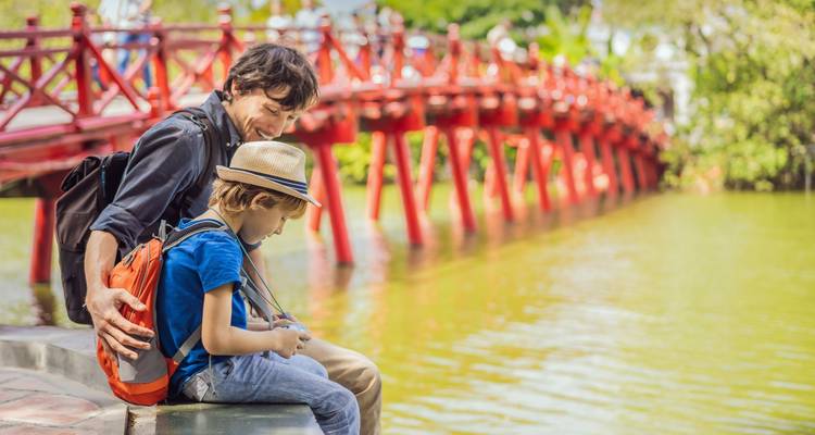 Un hombre y un niño se sientan junto a un cuerpo de agua con un puente rojo en el fondo.