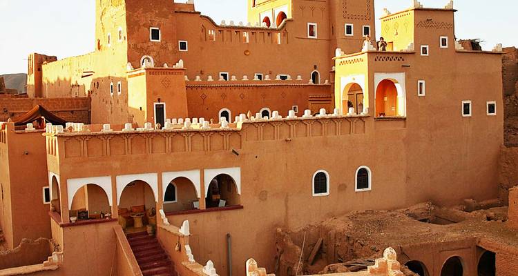 Kasbah de muros de adobe iluminada por el sol con parapetos almenados, ventanas arqueadas y paisaje desértico de fondo en Marruecos.