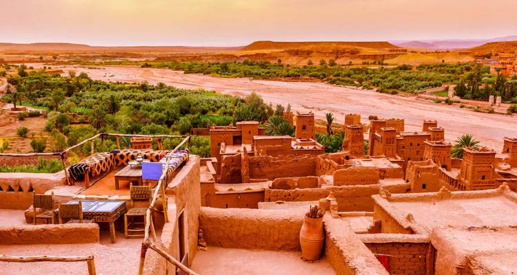 Vista panorámica del atardecer sobre un oasis exuberante y torres de kasbah de tierra con llanuras desérticas y mesetas más allá.