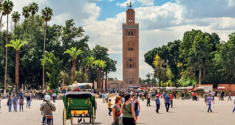 Plaza bulliciosa frente al minarete de la Mezquita de Kutubía con multitudes, carruajes de caballos y palmeras bajo un cielo brillante.