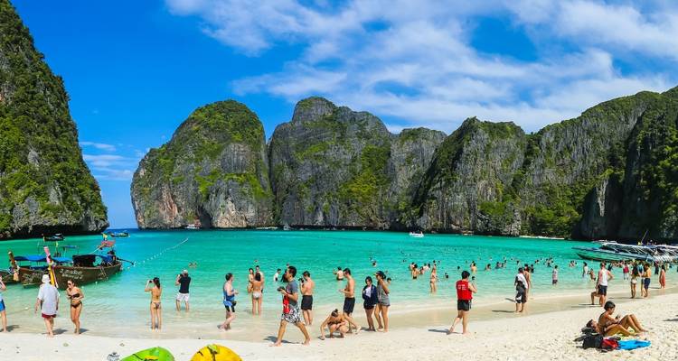 Menschenmengen von Urlaubern schwimmen und sonnen sich an einem Strand mit weißem Sand, der von steilen grünen Kalksteinfelsen und leuchtend türkisblauem Wasser umgeben ist.