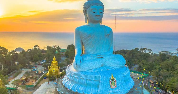 Eine riesige weiße Buddhastatue sitzt auf einem Hügel, der das Meer bei Sonnenaufgang überblickt, mit einer kleineren goldenen Buddha-Statue darunter und einem pastellfarbenen Himmel dahinter.