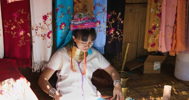 Mujer Karen tejiendo bufandas coloridas dentro de un puesto de madera tradicional, usando anillos de latón en el cuello.