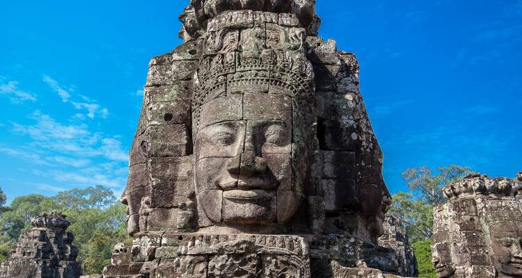 Reuze grijzende stenen gezichtstoren van Bayon Temple tegen een heldere blauwe lucht met omringende jungle.