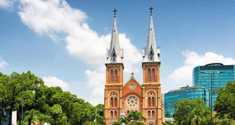 La Cathédrale Notre-Dame de Saigon aux briques rouges avec ses deux flèches encadrées par une végétation luxuriante et un ciel bleu.