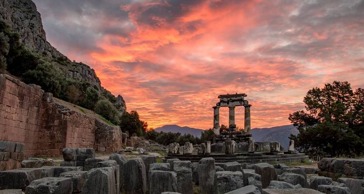 Ciel d'aurore embrasé au-dessus des ruines antiques de la Tholos à Delphes entourées de pierres rugueuses.