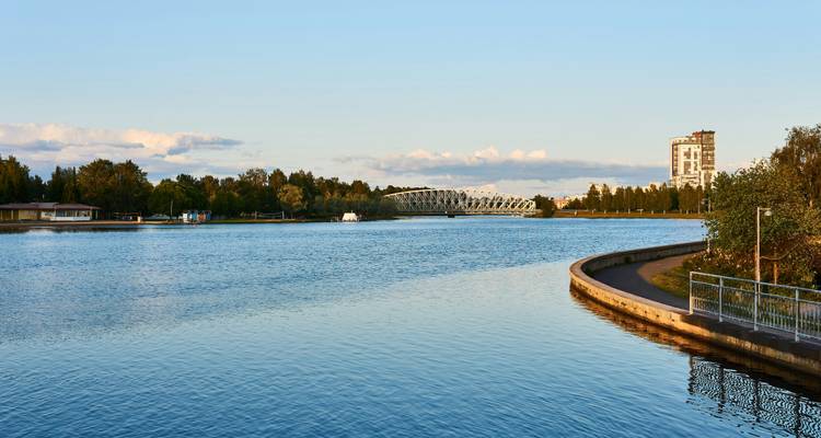 Ruhiger Fluss mit einer Brücke und Gebäuden in der Ferne unter einem klaren Himmel.