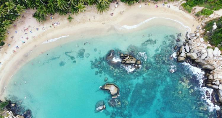 Vista de dron de una bahía turquesa, rocas de coral y playa dorada bordeada de palmeras exuberantes en el Parque Nacional Tayrona.