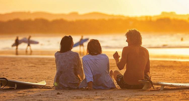 Tres surfistas se sientan en la arena con sus tablas mientras un atardecer dorado baña una playa australiana con luz cálida.