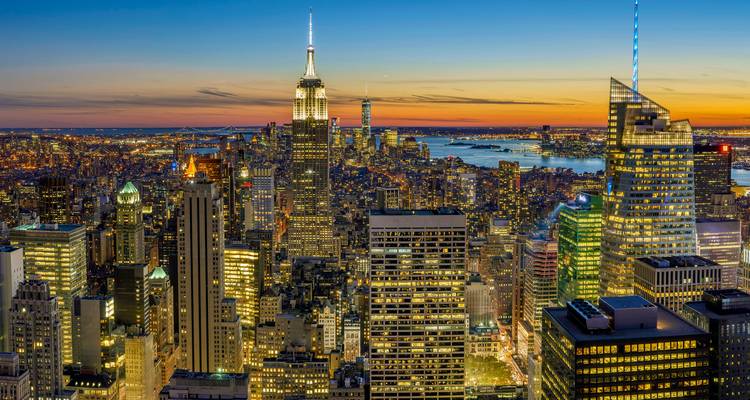 Panorama spectaculaire du coucher de soleil de la ville de New York avec les gratte-ciel illuminés, notamment l'Empire State Building et la lueur de l'Hudson River.