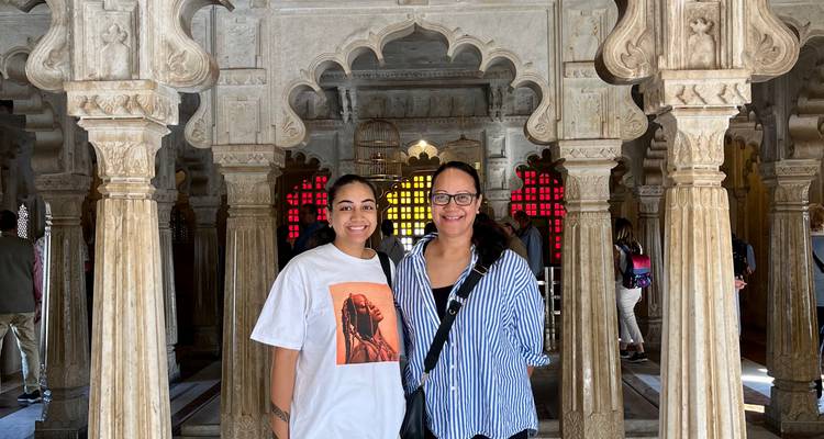Dos viajeros sonrientes posan dentro de un pabellón de mármol ornamentado con columnas talladas y ventanas de vidrieras coloridas en India.