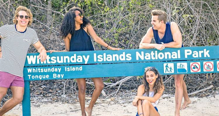 Quatre amis posent à côté d'un panneau du parc national des îles Whitsunday sur une plage de sable.