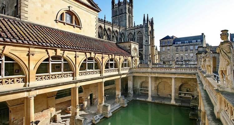 Historischer römischer Badekomplex mit dampfendem grünem Becken, das von goldenen Steinkolonnaden umrahmt wird, und der Bath Abbey dahinter.