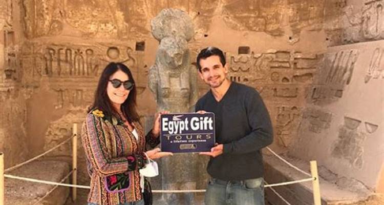 Pareja sonríe sosteniendo un cartel de 'Egypt Gift Tours' dentro de una cámara adornada con deidades talladas.