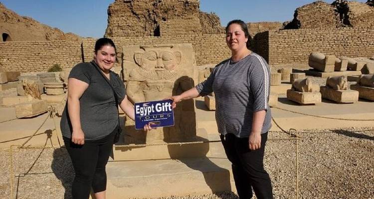 Deux voyageurs posent avec un panneau 'Egypt Gift Tours' au milieu de ruines de grès et de statues-blocs.