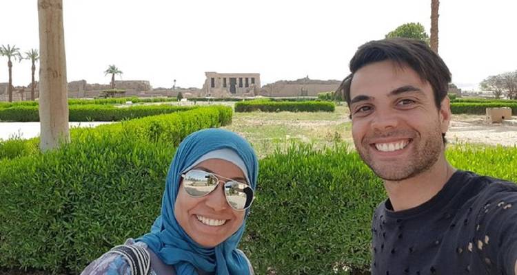Guide souriant et visiteur prennent un selfie avec des jardins luxuriants et des ruines de temple en arrière-plan.