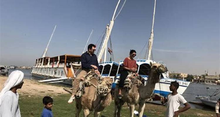 Des gens à dos de chameaux près du Nil avec des bateaux en arrière-plan.