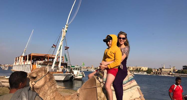 Femme et enfant montant un chameau près du fleuve Nil avec des bateaux en arrière-plan.