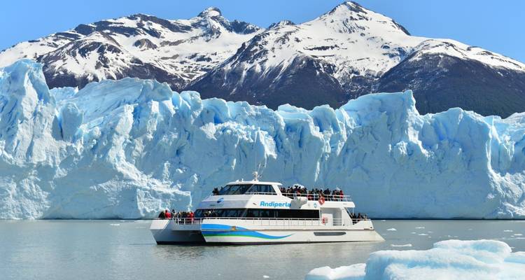 Ein Boot, das vor einem großen Gletscher fährt, mit schneebedeckten Bergen im Hintergrund.