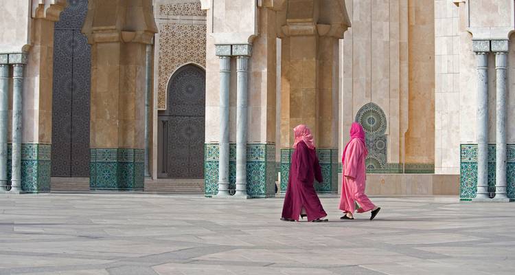 Dos mujeres con túnicas vibrantes caminan por el vasto patio de mármol de una gran mezquita adornada con intrincados mosaicos.