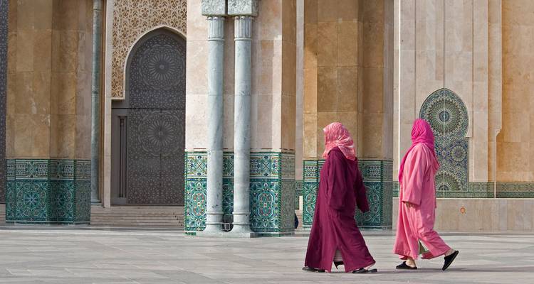 Two women in colorful djellabas walk past intricate tilework of a grand Moroccan mosque.