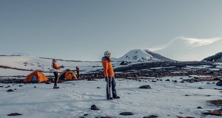 Petit camp d'expédition aux tentes oranges installé sur un plateau enneigé face à un pic volcanique à l'aube.