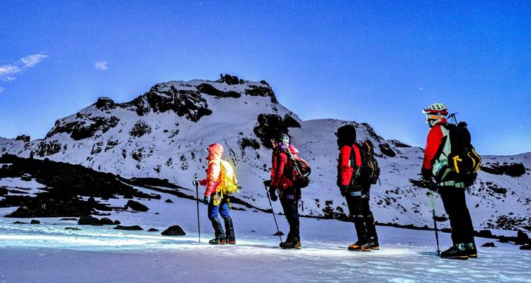 Des alpinistes équipés de lampes frontales s'alignent sur un champ de neige luminescent au pied d'un pic rocheux avant l'aube.
