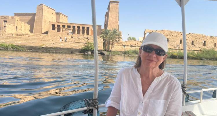 Frau in Weiß sitzt auf einem Boot mit dem Philae-Tempel und Palmen im Hintergrund.