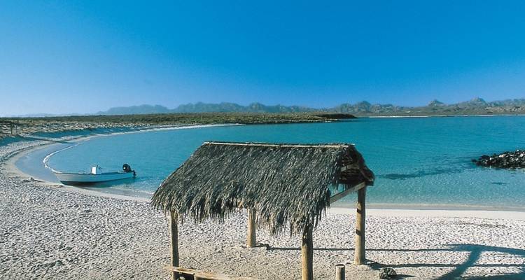 Eine kleine Hütte mit Strohdach an einem Sandstrand mit einem Boot im Wasser und Bergen in der Ferne.