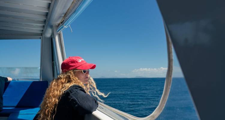 Femme au bonnet rouge contemple la mer d'un bleu profond depuis le bastingage d'un bateau rapide sous un ciel dégagé.
