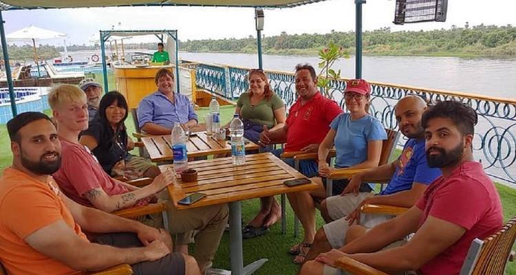 Travelers relax around a table on the upper deck of a Nile cruise boat with the river and palm-lined shore behind them.