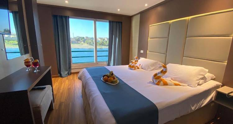 Modern cabin with a large bed, towel swans and fruit plate, featuring a panoramic river view through floor-to-ceiling windows.