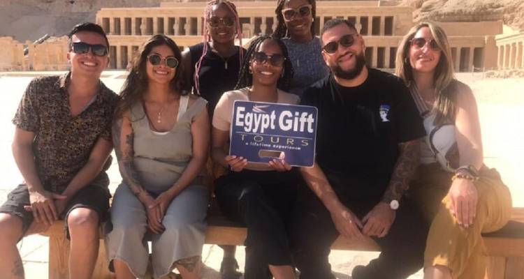 Seven travelers sit on a bench holding an 'Egypt Gift Tours' sign with the Temple of Hatshepsut rising behind them.