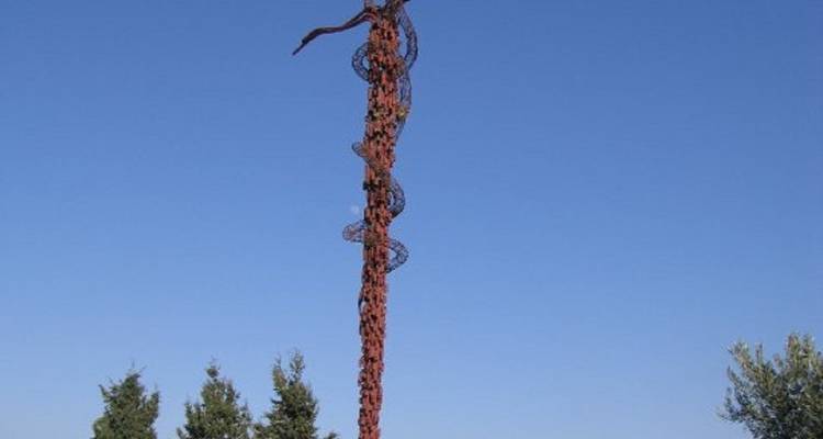 Hoge metalen sculptuur die lijkt op een slang op een kruis met bomen op de achtergrond.