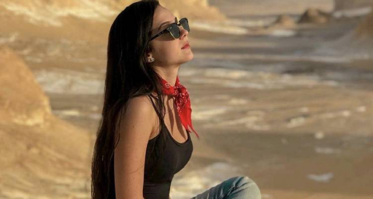 Woman in sunglasses and red scarf sits on desert ground admiring sunlit chalk formations.