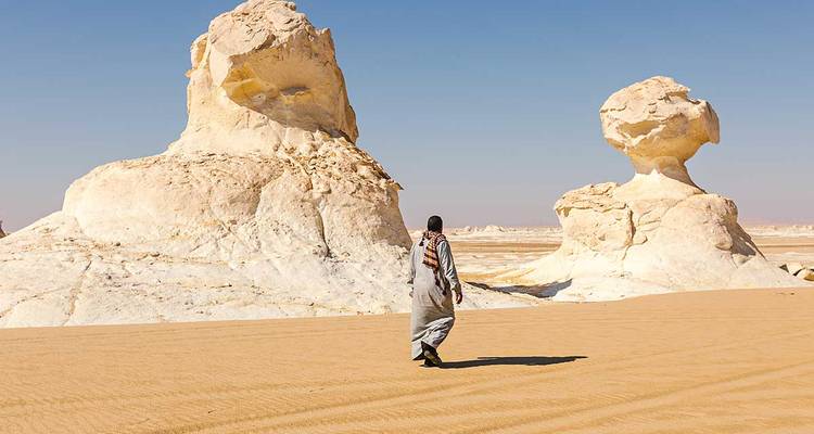 Traveler in traditional robe walks toward surreal mushroom-shaped white rock formations in a sandy desert.