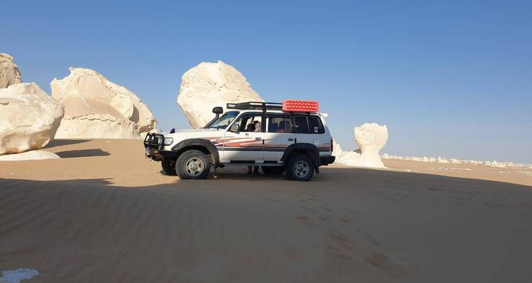 SUV parked on rippled desert sand with white rock pillars rising behind under blue sky.