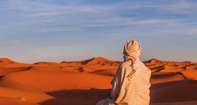 A lone traveler wrapped in traditional cloth sits quietly atop vivid orange desert dunes under a pastel sky.