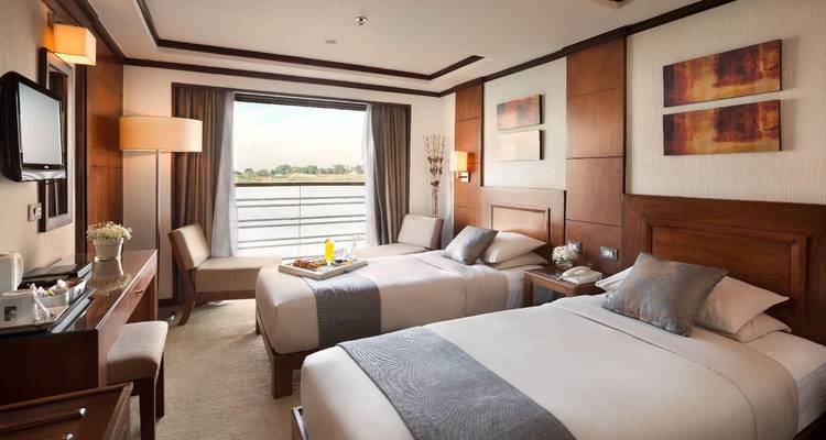 Camarote doble moderno y luminoso con ventana con vista al río a bordo de un crucero por el Nilo, ordenadamente dispuesto con decoración neutra.